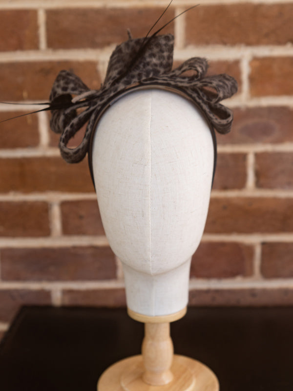 Front view of leopard printed felt headpiece. There is a felt bow design on top with thin feather trimming.