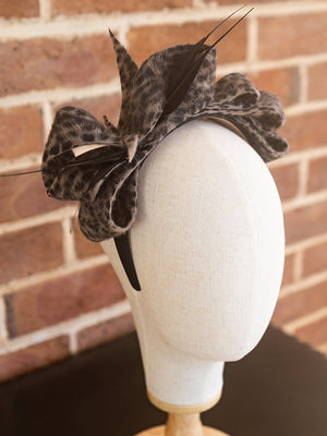 Side view of leopard printed felt headpiece. There is a felt bow design on top with thin feather trimming.